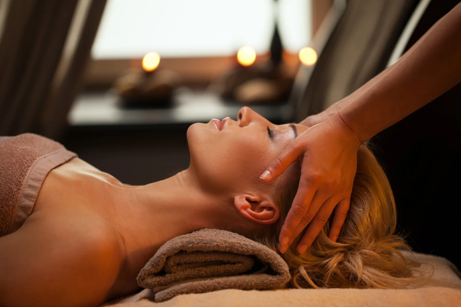 A client's neck supported by a therapist's hands, with a rolled towel and candlelight nearby.